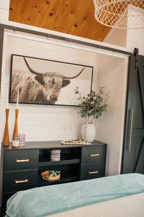 Une chambre à coucher avec un lit, une commode à tiroirs, un tableau encadré au mur, un vase avec une plante et un meuble en bois.