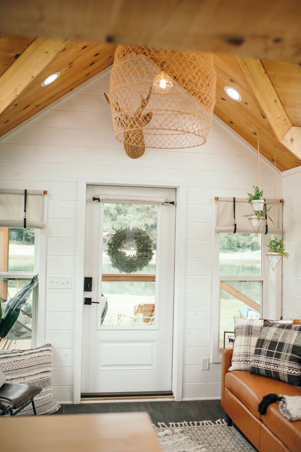 Un intérieur chaleureux avec un plafond en bois, une porte blanche ornée d’une guirlande, un canapé en cuir marron et un luminaire suspendu.