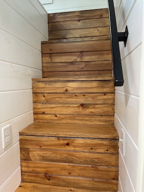 Un escalier en bois avec une rampe noire, des murs blancs et une prise électrique visible.