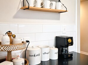 A kitchen with white tiles, a coffee maker, and labeled storage containers for cookies, tea, and coffee.