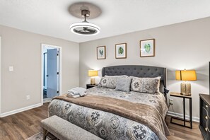 A bedroom with a large bed, two bedside lamps, a ceiling fan, and a door leading to another room.