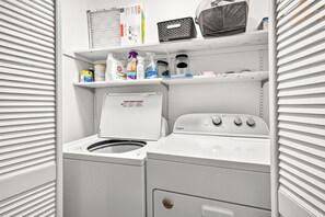 Une petite buanderie équipée d’un lave-linge et d’un sèche-linge à chargement frontal, d’étagères contenant des produits de nettoyage et d’un petit four à micro-ondes posé sur le plan de travail.