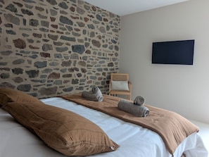 A bedroom with a stone wall, a bed with brown bedding, a wooden chair, and a dark framed picture on the wall. - L'Atelier - Maison avec piscine face au volcan (Saint-Just-près-Brioude)