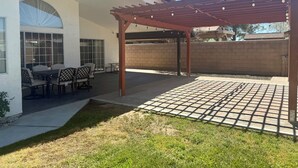 Um alpendre coberto com mesa e cadeiras, uma pérgula com luzes de cordão e um muro de tijolos ao fundo.