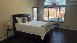 Quarto com uma cama grande, mesa de cabeceira, luminária, janela com persianas e ar condicionado.