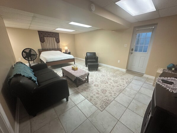 A bedroom with a bed, a bedside table with a lamp, a fan, a leather sofa, a small table with a decorative item, and a door with a window.