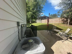 Un patio con ventilador, mobiliario exterior y vista a un jardín con valla de madera y árboles.