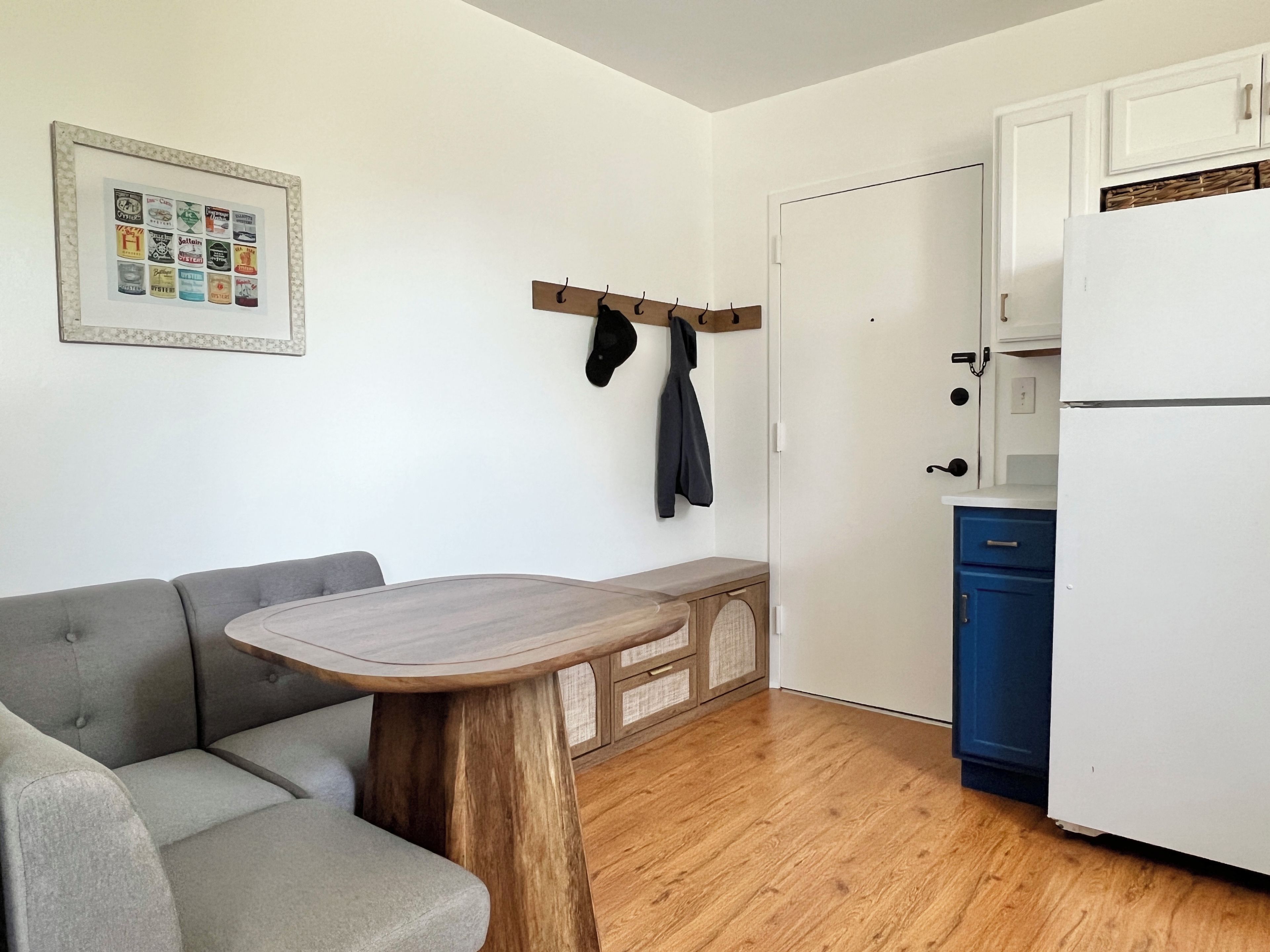 Un espacio habitable compacto con un sofá gris, una mesa de madera, una zona de cocina con un refrigerador blanco y una puerta con perchero.