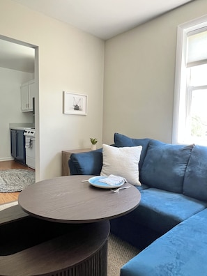 A modern living room with a blue sofa, a round coffee table, and a kitchen area visible in the background.