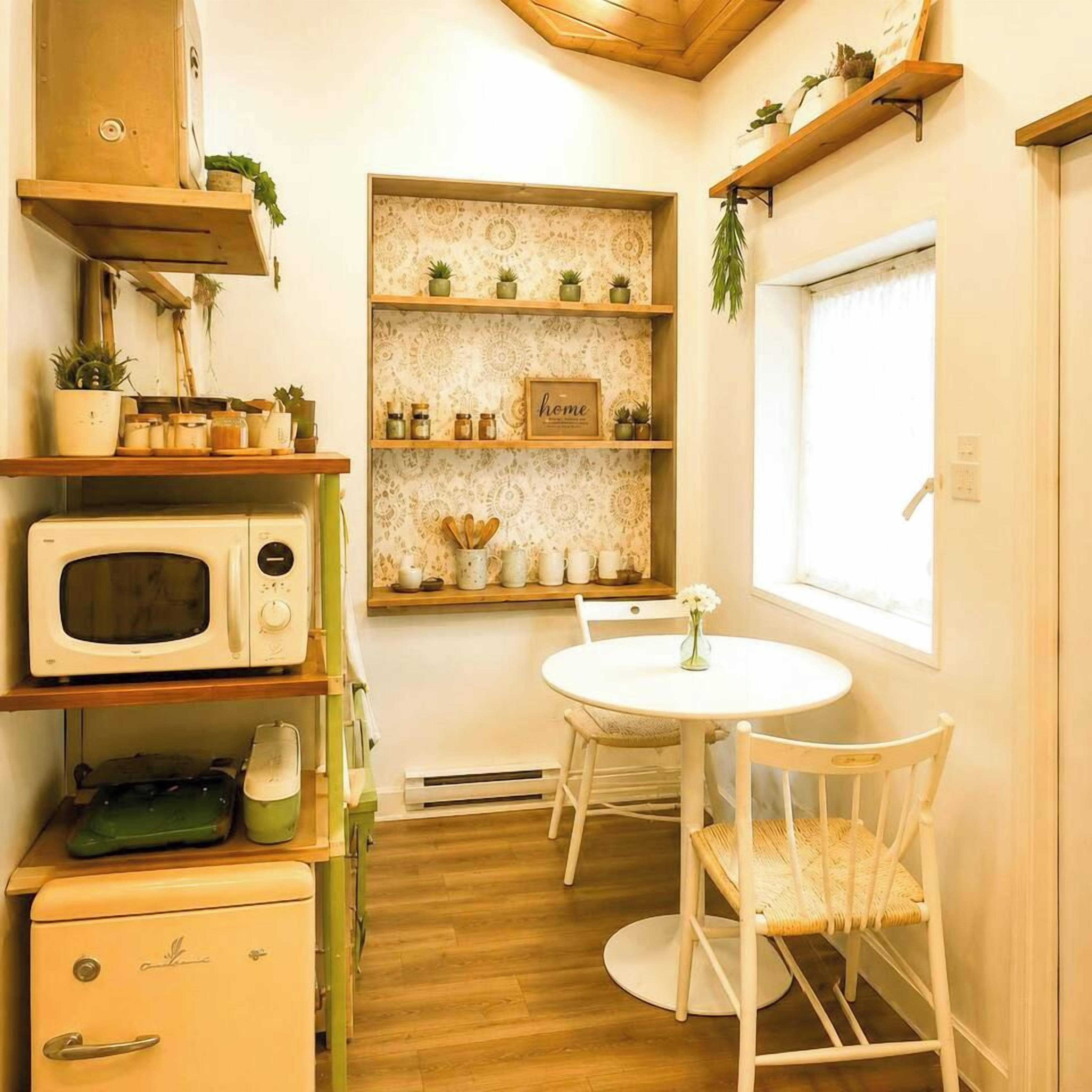 Een praktische keuken met een magnetron, een kleine koelkast en een eettafel met stoelen. Er staan planken met planten en keukengerei.