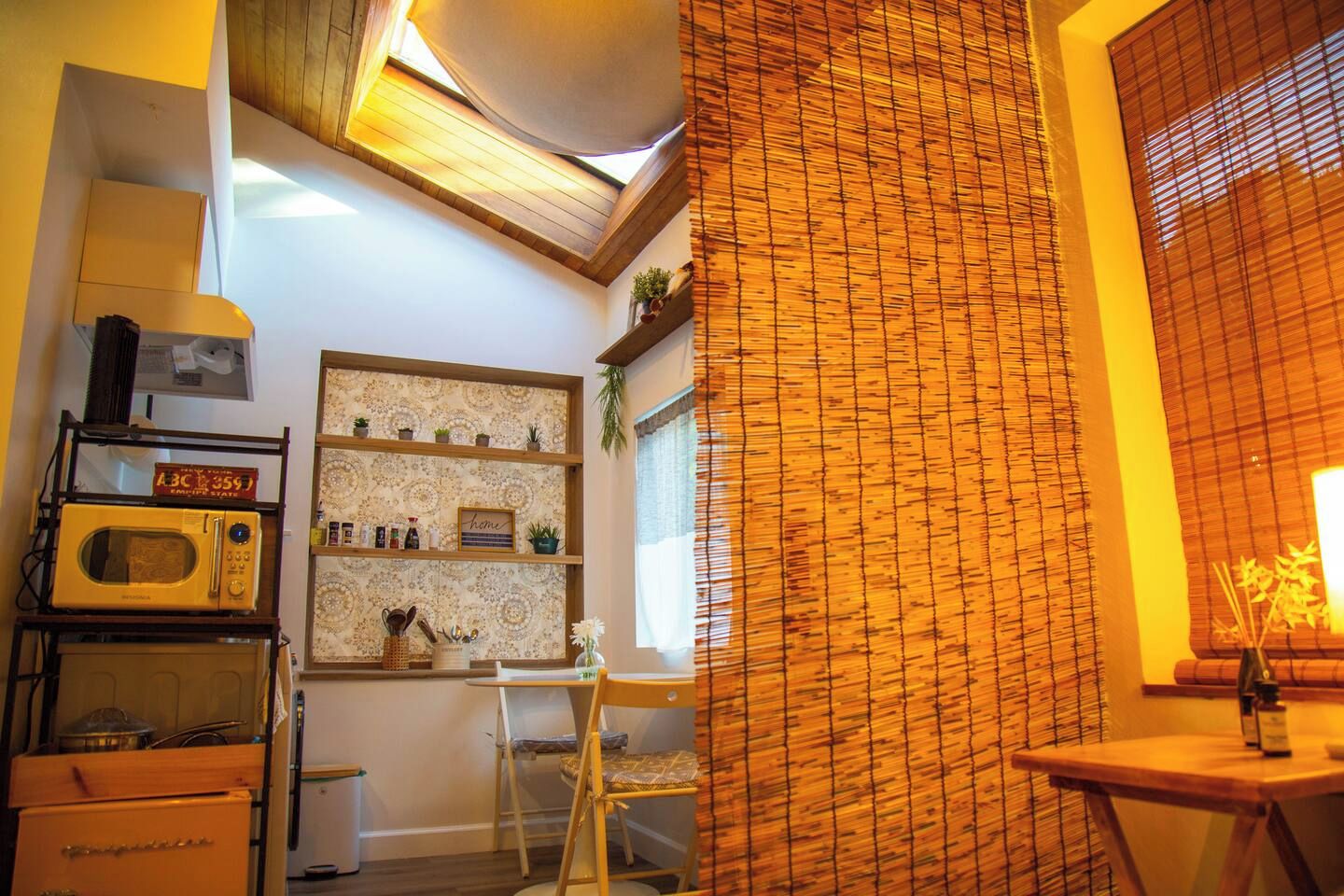 Eine Küche mit Mikrowelle, Regalen voller Küchenutensilien, einem Fenster mit Jalousien und einem Holzstuhl.