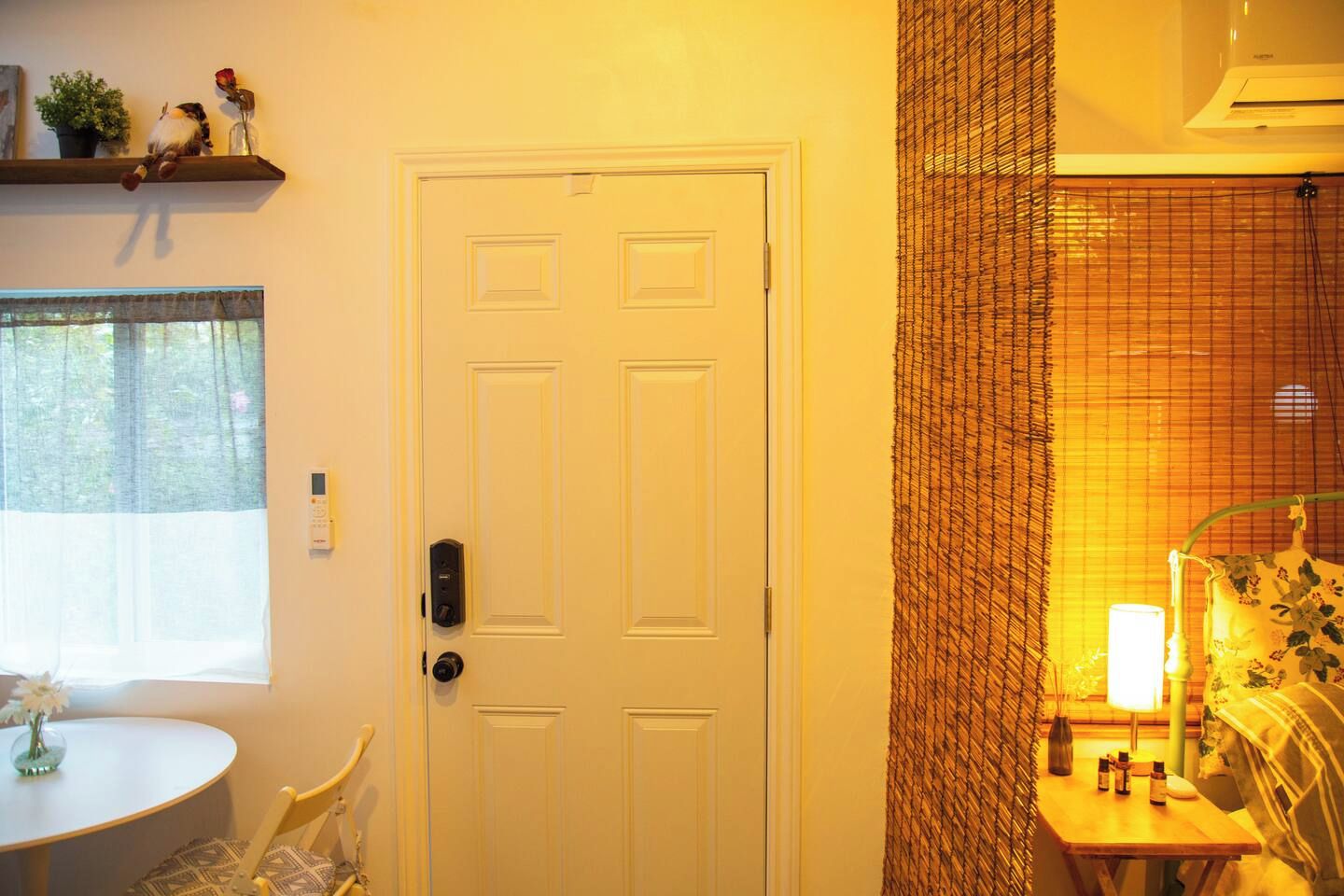 Una habitación con una puerta blanca, una mesita con un florero y una ventana con cortinas translúcidas.