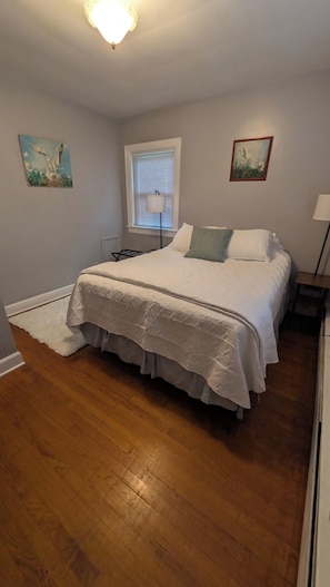 A bedroom with a bed, a window with blinds, a painting on the wall, and a lamp beside the bed.