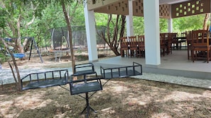 An outdoor seating area with tables and chairs, surrounded by trees and a swing set.