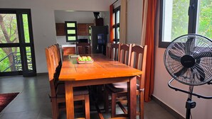 A dining area with a wooden table, chairs, and a fruit basket. There is a large window with curtains and a standing fan. - Suñu Tool Private Farm Guesthouse, ferme agroécologique, entourée de nature. (Thiès)