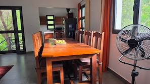 A dining area with a wooden table, chairs, and a fruit basket. There is a large window with curtains and a standing fan.