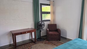 Una habitación con un escritorio de madera, una silla, un ventilador de pie, una ventana con cortinas y una alfombra estampada.