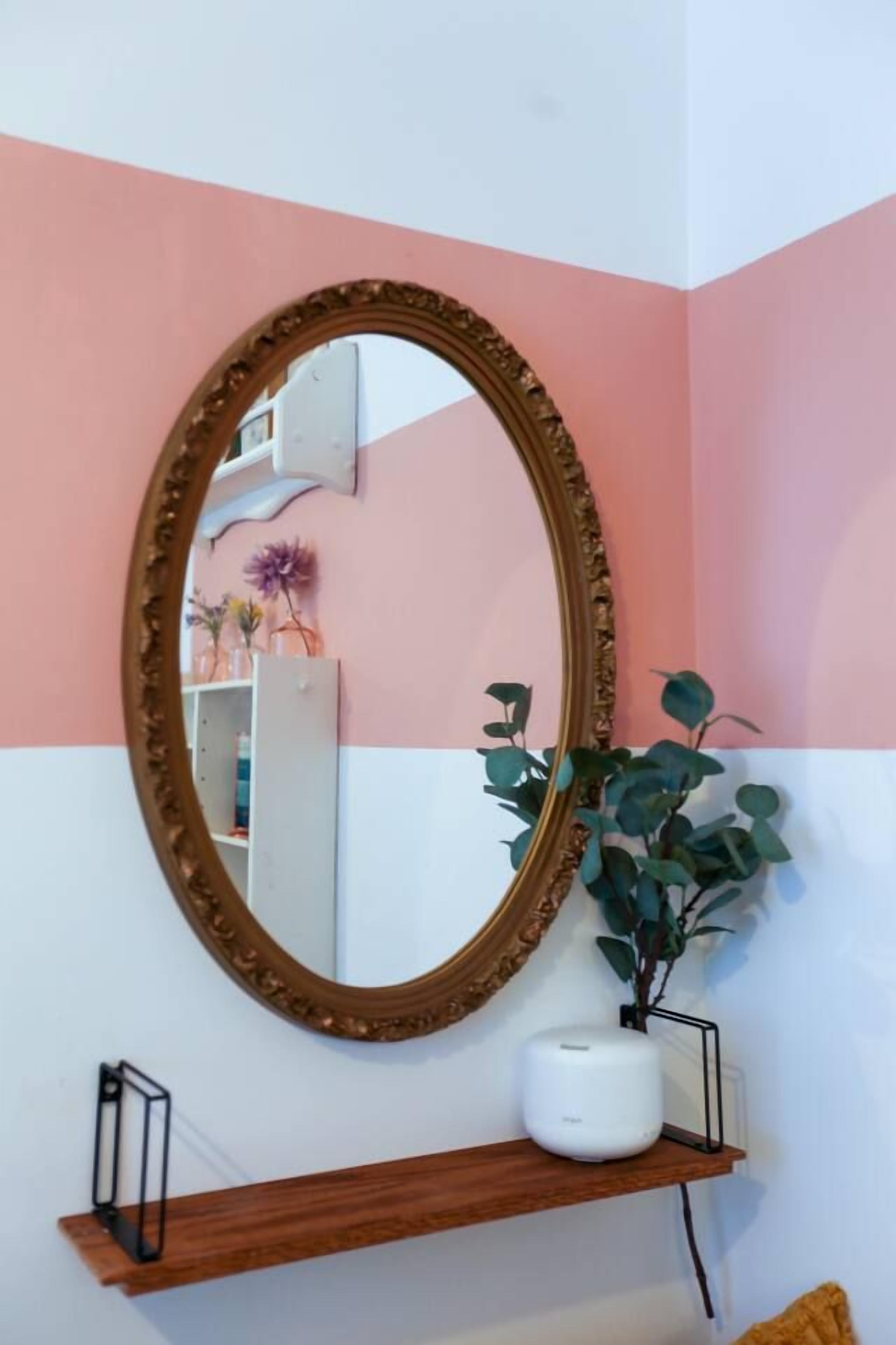 Een spiegel met een sierlijk kadertje aan een muur, een potplant en een plankje met een wit voorwerp.