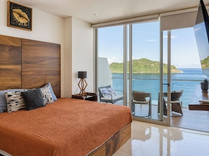 Ein Schlafzimmer mit einem großen Bett, einem Nachttisch, einer Stehlampe und Blick auf das Meer durch eine Schiebetür.