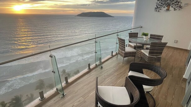 A balcony with a glass railing overlooking the sea, furnished with wicker chairs and a table. - Beachfront Luxury | Camino al Mar (Mazatlán)