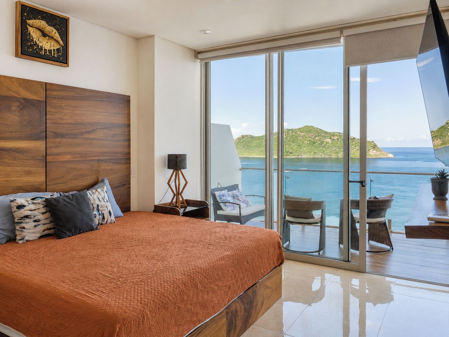 A bedroom with a large bed, a nightstand, a floor lamp, and a view of the sea through a sliding glass door.