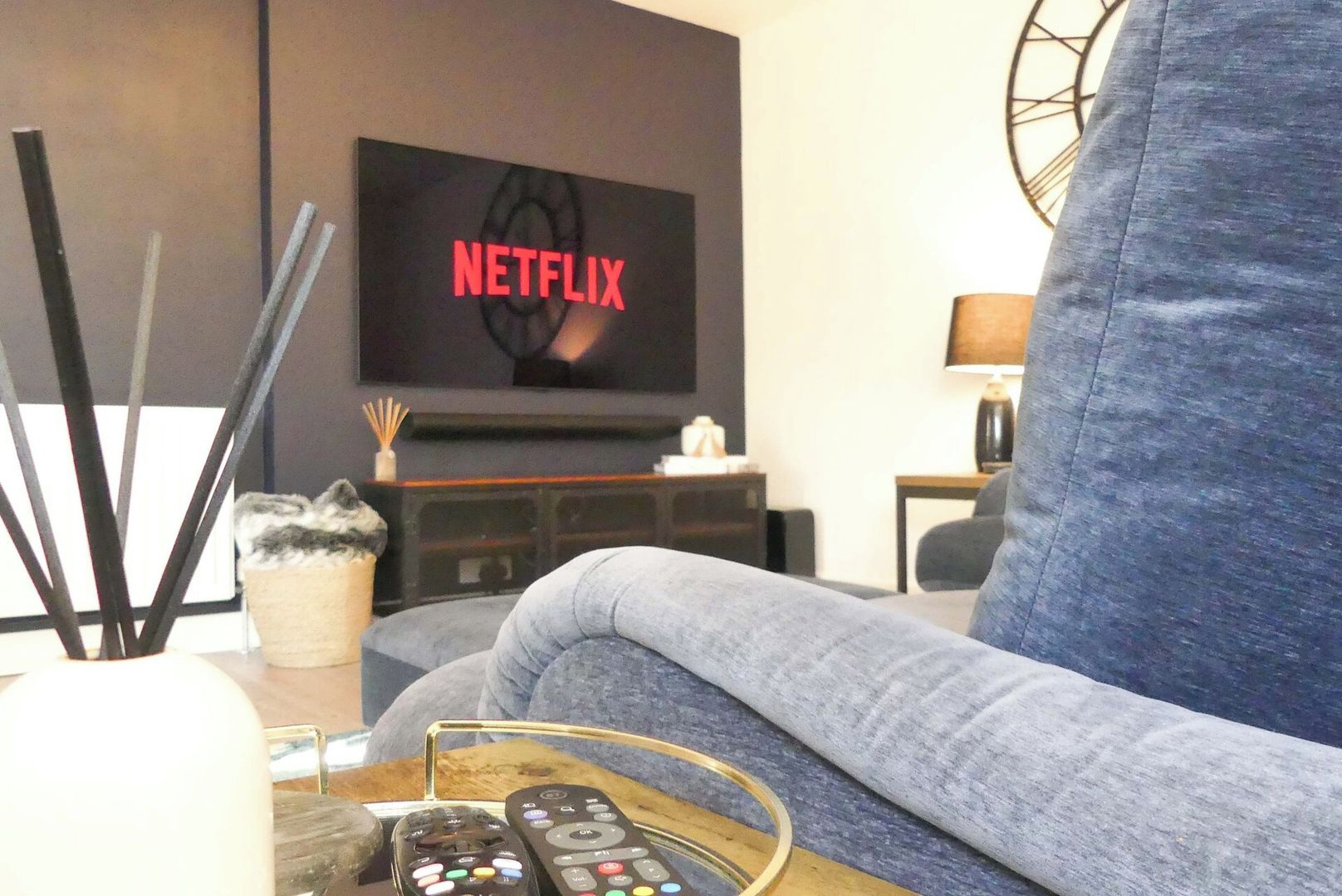 A living room with a grey sofa, a coffee table with remote controls, and a TV displaying the Netflix logo.