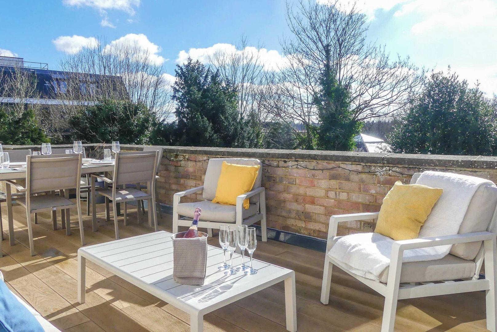An outdoor patio with white furniture, including chairs and a table, set against a brick wall with a view of trees and a building in the background.