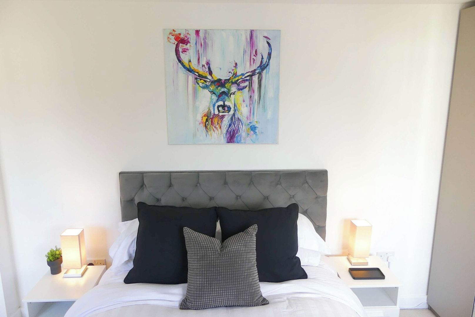 Une chambre à coucher avec une tête de lit capitonnée, une literie blanche, des oreillers noirs et gris, des lampes de chevet et une peinture abstraite colorée accrochée au-dessus du lit.