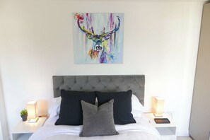 A bedroom with a tufted headboard, white bedding, black and grey pillows, bedside lamps, and a colorful abstract painting above the bed.