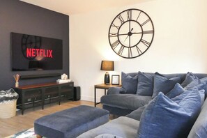 Olohuoneessa on suuri taulutelevisio, moderni kello, sohva ja pieni pöytä, jossa on lamppu.
