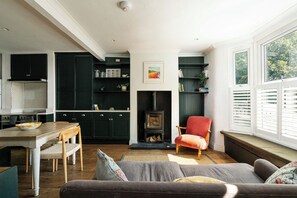 A cozy living room with a fireplace, a dining area with a wooden table, and a kitchen with dark green cabinets. - Comfortable 3BR Townhouse for 6 (London)