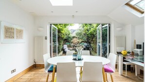 Espaço de refeições moderno com mesa redonda e cadeiras, vista para um jardim através de portas de vidro, e decoração em tons brancos com claraboia.