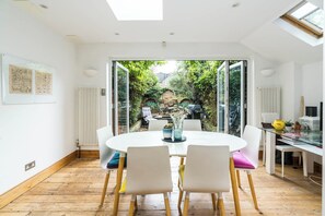 A modern dining area with a round table and chairs, a view of a garden through glass doors, and a white interior with a skylight. - Stunning 4 Bedroom Home w/Garden in Fulham (Greater London)