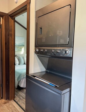 A compact washer-dryer unit with a control panel, located in a room with a wooden door and a bed with white bedding. - Tiny living, fire pit, pond fishing!!! (Boaz)