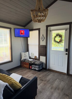 A room with a television, a bench with storage, a door with a wreath, and a chandelier. - Tiny living, fire pit, pond fishing!!! (Boaz)