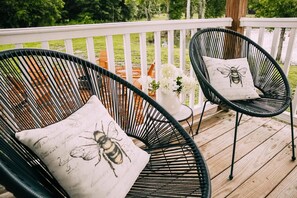 Deux fauteuils en osier pour l’extérieur, dotés de coussins à motifs d’abeilles, sont placés sur une terrasse en bois avec une rambarde blanche.