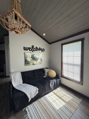 Habitación acogedora con un sofá de color oscuro, un letrero de bienvenida, un cuadro y un ventanal con persianas.