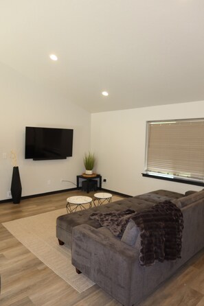 A modern living room with a grey sofa, a flat-screen TV mounted on the wall, a small table with a plant, and a window with blinds. - Charming 3-bedroom house in serene Athol! 5 Min to Silverwood. Firepit (Athol)
