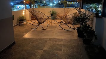 Un espace extérieur aménagé en terrasse, avec un sol en pierre, deux fauteuils à bascule, des plantes en pot et des guirlandes lumineuses.