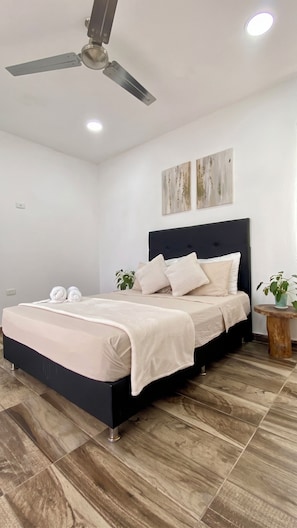 Una camera da letto con un letto, una ventola a soffitto, opere d'arte alle pareti e un piccolo tavolino con una pianta.