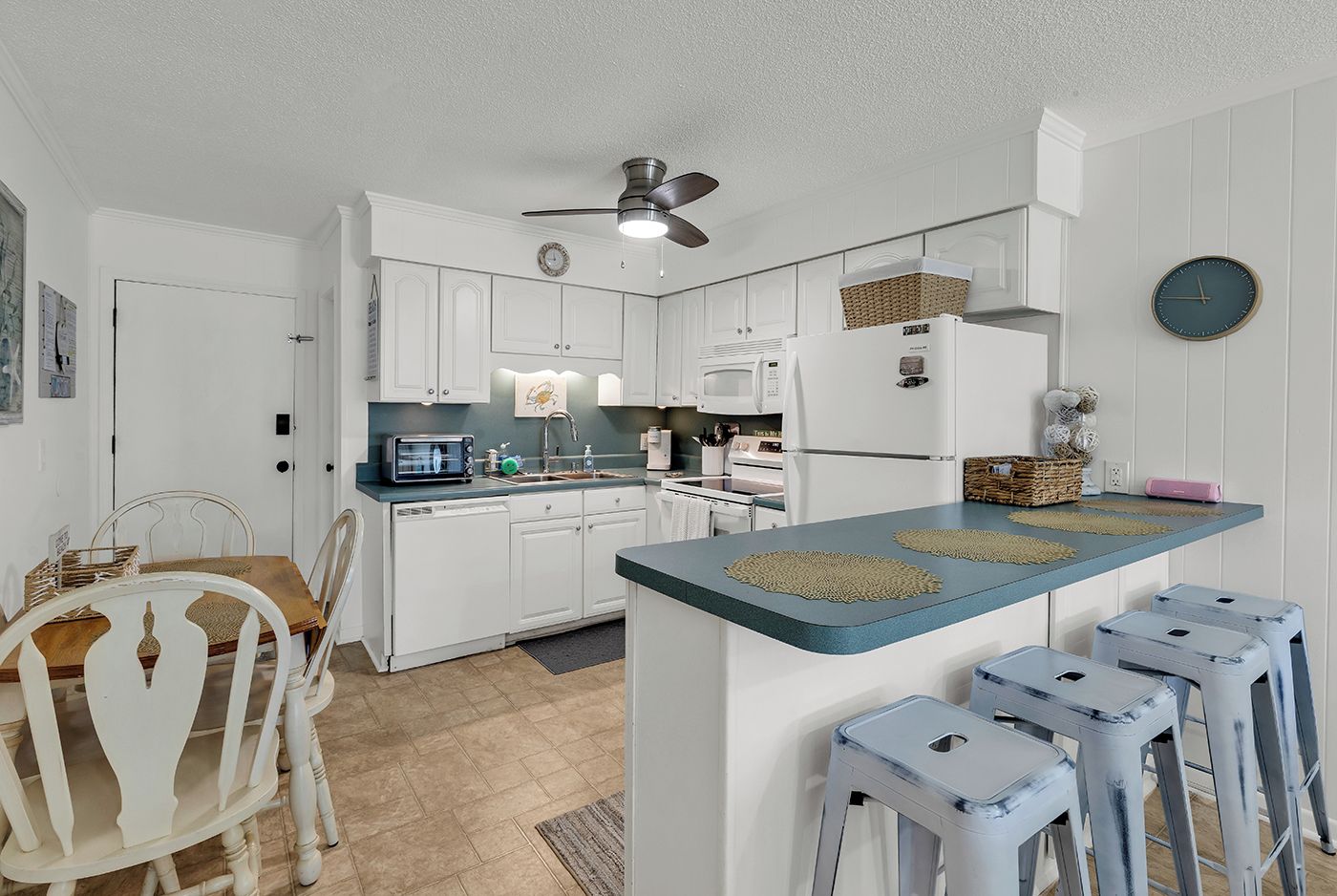 A compact kitchen with white cabinetry, a blue countertop, a ceiling fan, and a clock on the wall.