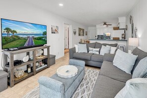 Een moderne woonkamer met een flatscreen televisie, grijze banken en een keuken op de achtergrond.
