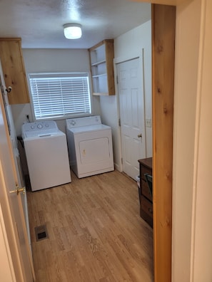 A laundry room with two white washing machines, a wooden cabinet, a white door, and a window with blinds.