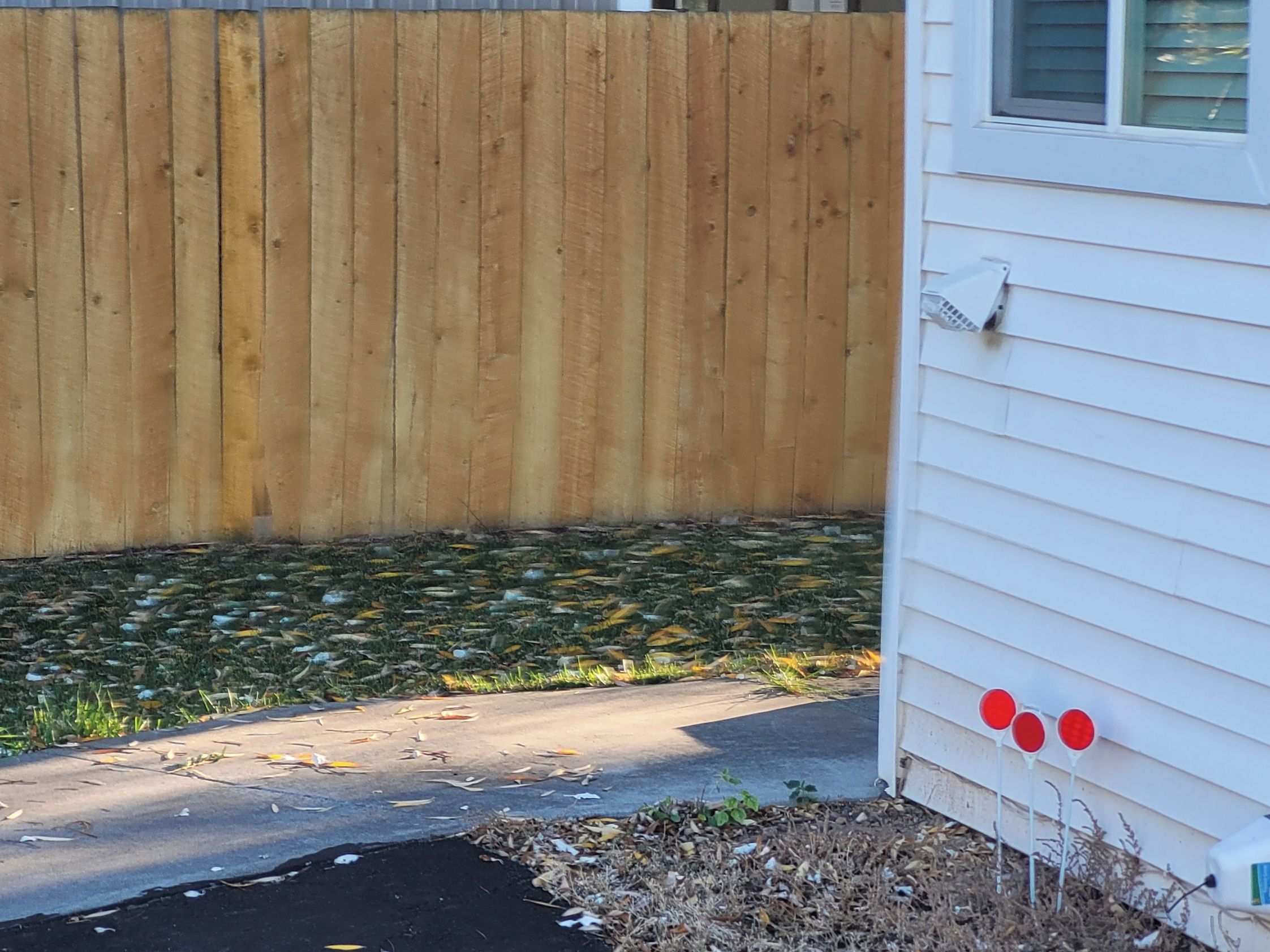 A corner of a house with a wooden fence, a concrete driveway, and a white exterior wall with three red lights.