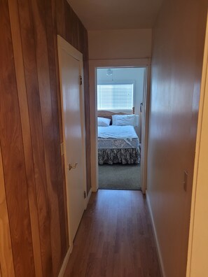 A narrow hallway leading to a bedroom with a bed, a window with blinds, and a door on the left. - White 2-bedroom house in perfect Mountain View location (Mountain View)