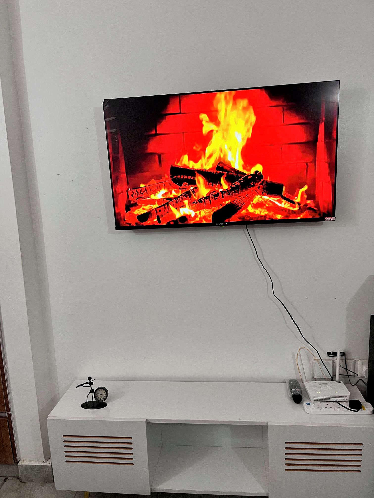 Ein Flachbildfernseher ist an einer weißen Wand angebracht und zeigt ein Lagerfeuer. Darunter befindet sich eine weiße Unterhaltungseinrichtung mit einem Lautsprecher und einem kleinen Dekorationsgegenstand.