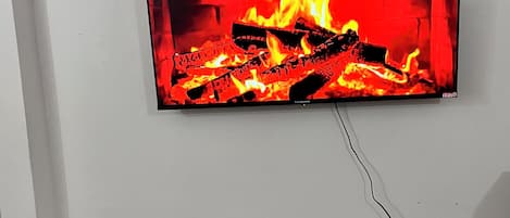 Televisão de tela plana presa a uma parede branca exibindo uma lareira. Abaixo da televisão, um móvel branco com um alto-falante e um pequeno objeto decorativo.