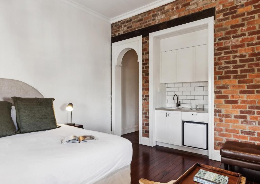 Un dormitorio con una cama grande, una mesita de noche con lámpara y vistas a una cocina con armarios blancos y una pared de ladrillo.