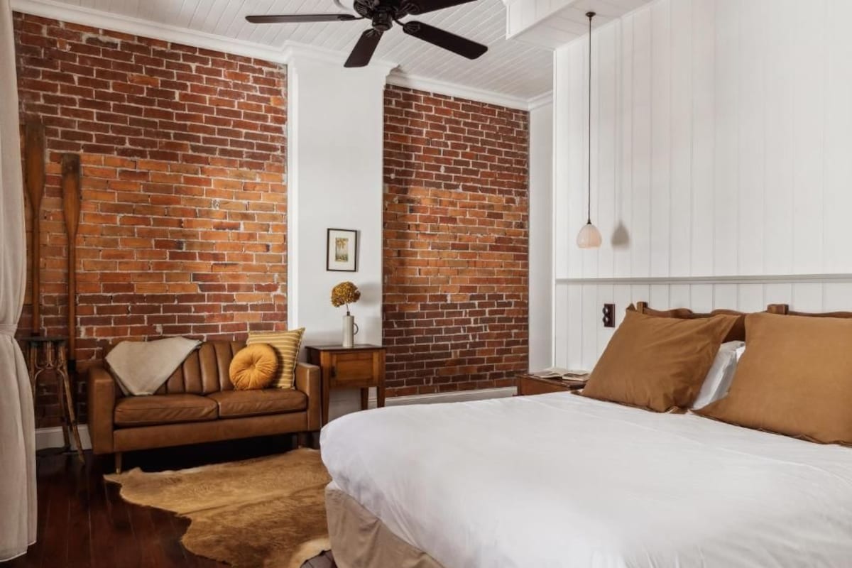 Quarto com parede de tijolos, cama com roupa de cama branca, sofá marrom e ventilador de teto.