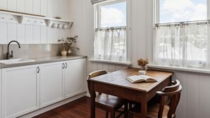 Cozinha com armários brancos, mesa de madeira e duas janelas com cortinas.